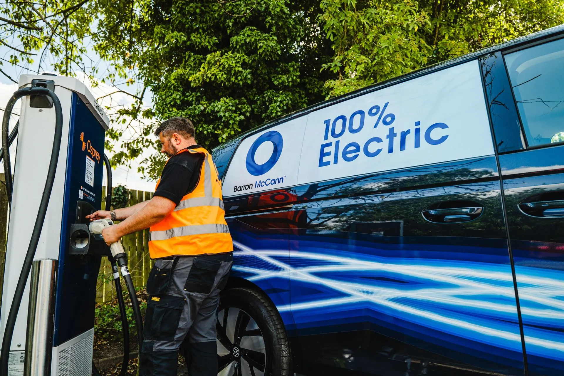 A Barron McCann engineer in a hi-vis vest using a rapid charger beside an electric Barron McCann van